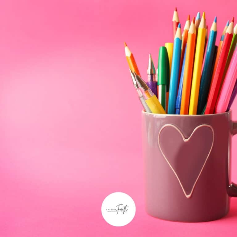 Pink mug with a heart design holding colorful pencils and pens against a pink background